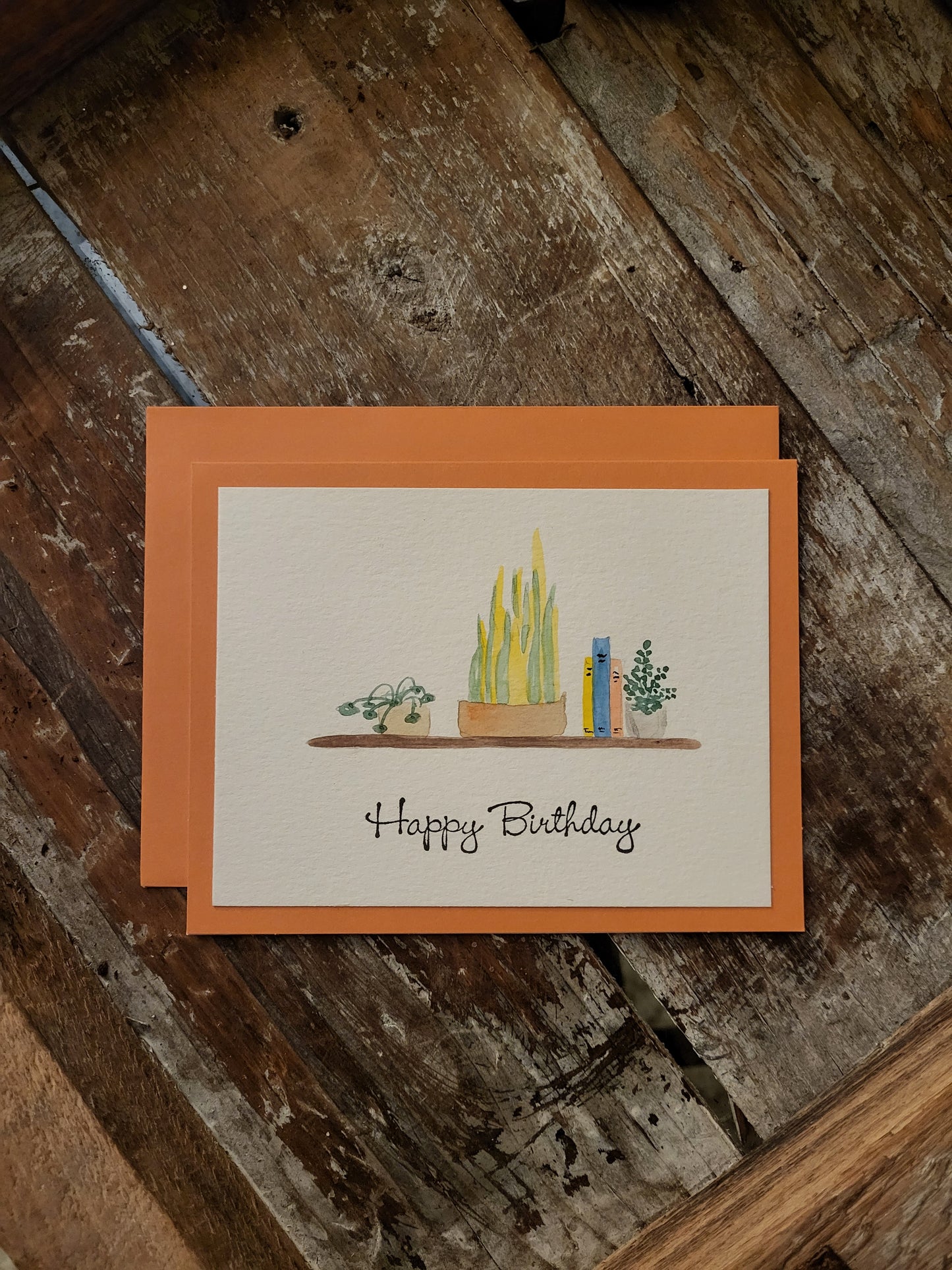 A collection of painted plants and books sit on a shelf. A stamp says Happy Birthday.