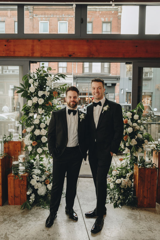 Two grooms stand in front of an asymmetrical ceremony installation with white and toffee flowers and foliage. 